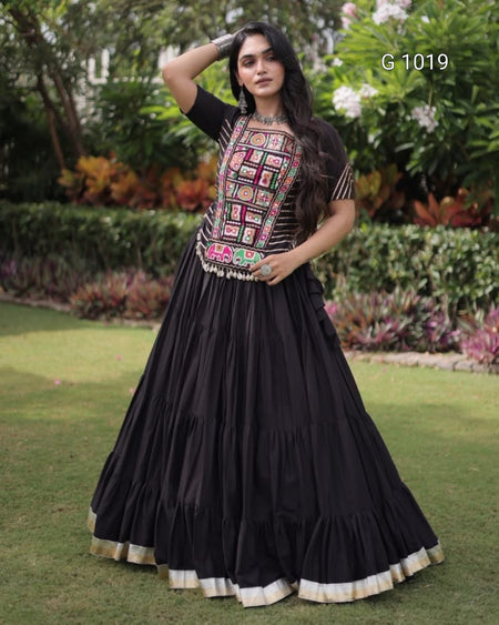 A woman modeling a black traditional dress with a heavily embroidered multicolor bodice and a tiered black skirt with a gold-trimmed hem, standing on a green lawn with garden foliage in the background.