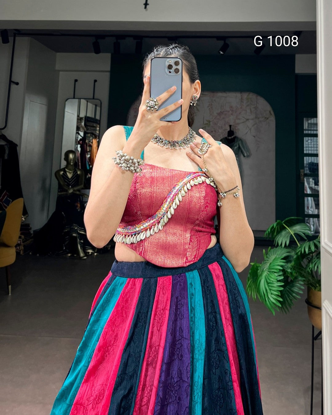 A woman takes a mirror selfie wearing a traditional Indian outfit with a metallic pink and teal crop top, a silver embellished waist chain, and a striped skirt in pink, teal, and purple. She is wearing heavy silver jewelry, including a choker necklace, bracelets, and rings.