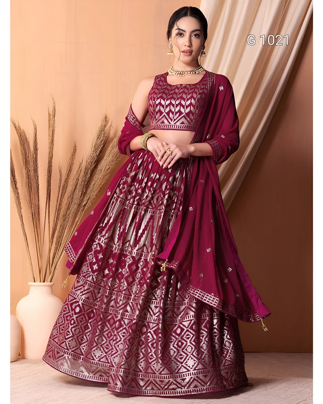 A woman models a deep magenta lehenga choli set with intricate silver mirror work embroidery, set against a warm beige background with draped curtains and a vase of dried grass.