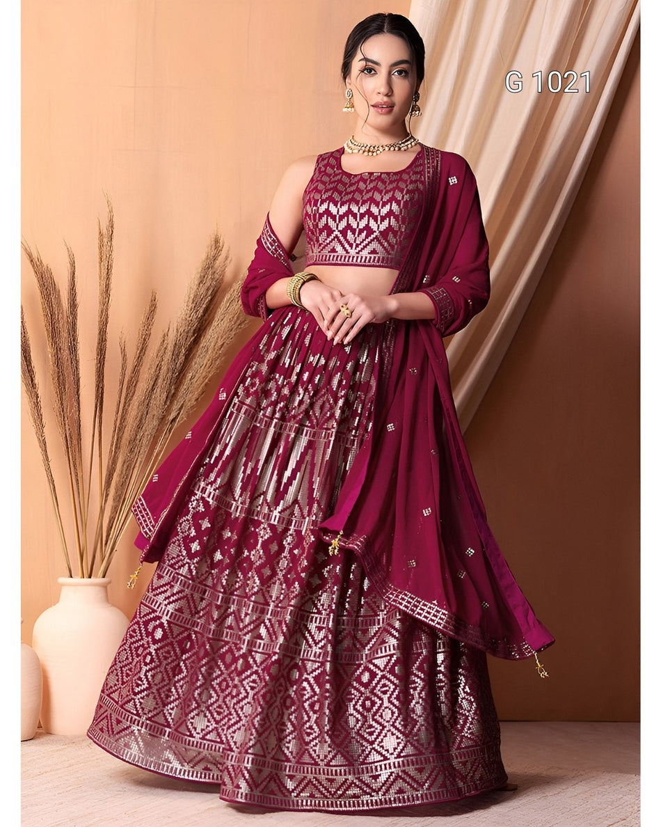 A woman models a deep magenta lehenga choli set with intricate silver mirror work embroidery, set against a warm beige background with draped curtains and a vase of dried grass.