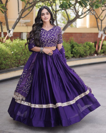 A woman modeling a purple traditional Indian lehenga choli set outdoors. The outfit features a heavily embroidered purple blouse with three-quarter sleeves, a matching pleated skirt, and a dupatta with silver mirror work and metallic border detailing.