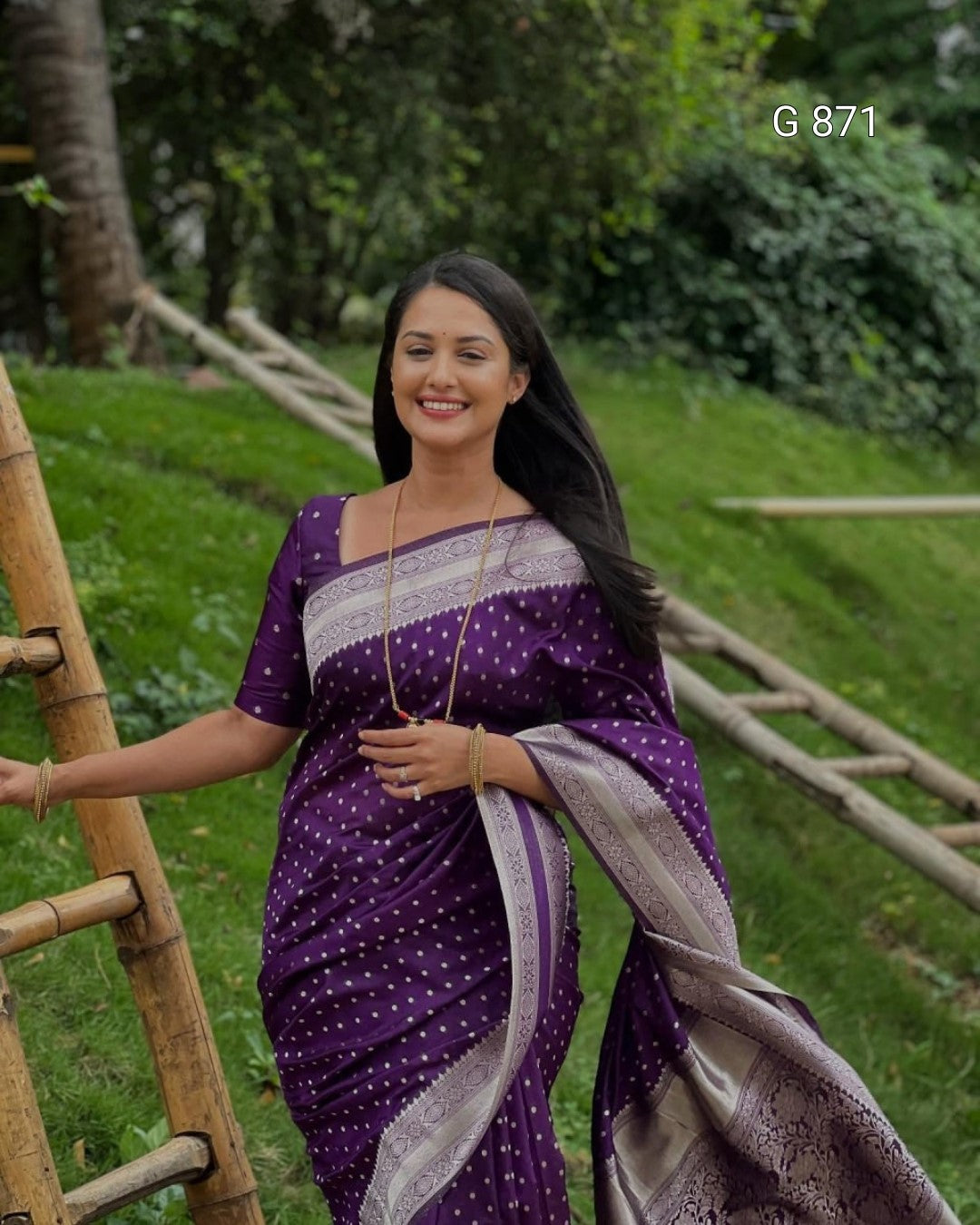 Woman in a purple saree with white patterns standing outdoors with greenery in the background