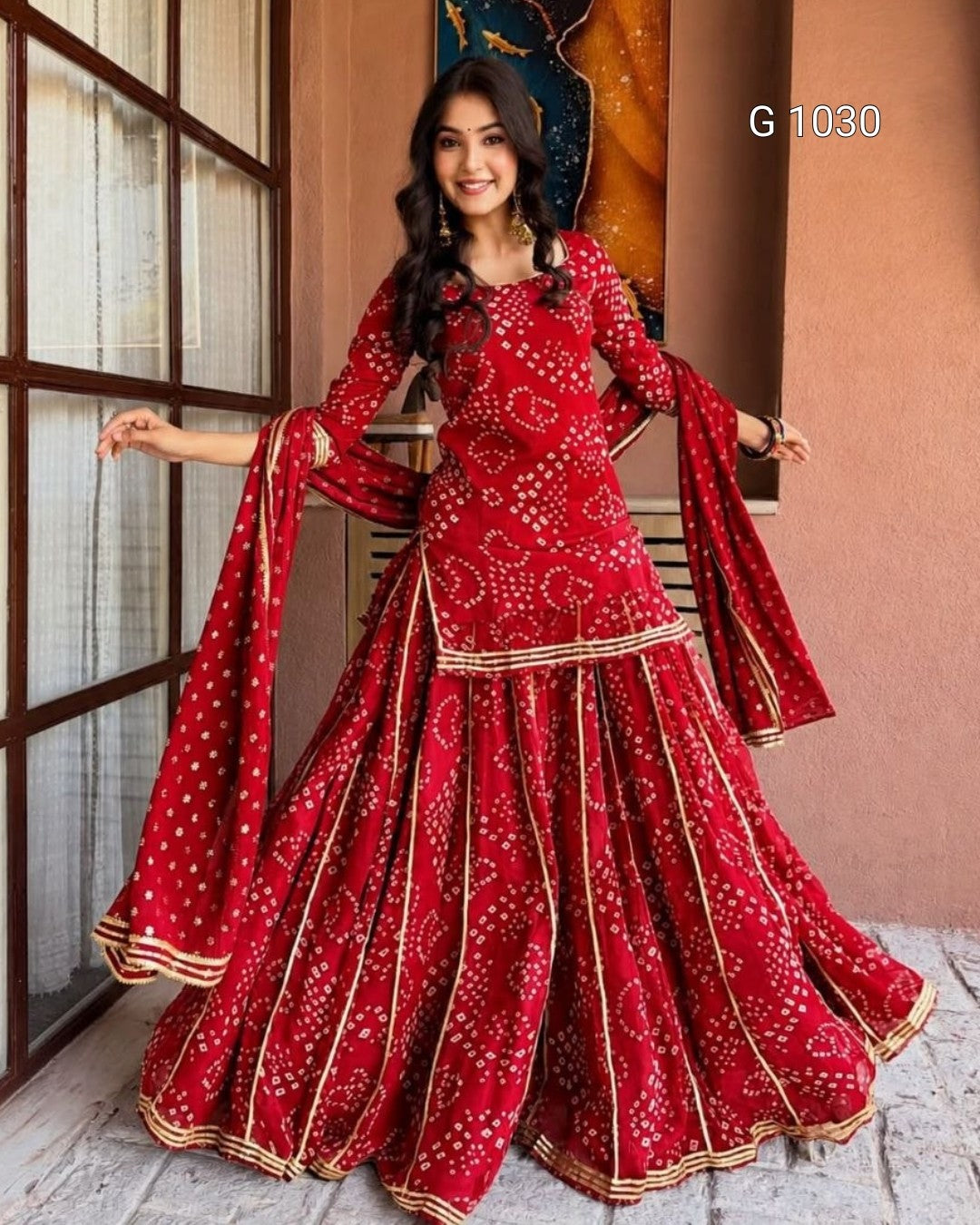 A woman modeling a red traditional Indian ethnic outfit with white bandhani print and gold borders, featuring a flared skirt and matching dupatta, posed in front of a rustic indoor backdrop.