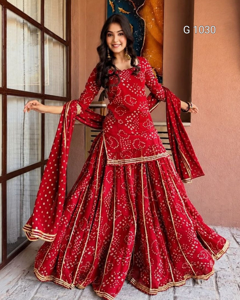 A woman modeling a red traditional Indian ethnic outfit with white bandhani print and gold borders, featuring a flared skirt and matching dupatta, posed in front of a rustic indoor backdrop.