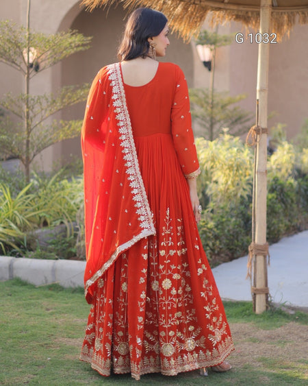 A woman wearing a red traditional ethnic outfit with gold embroidery and a matching dupatta, photographed outdoors on a lawn with plants and a pavilion structure in the background.