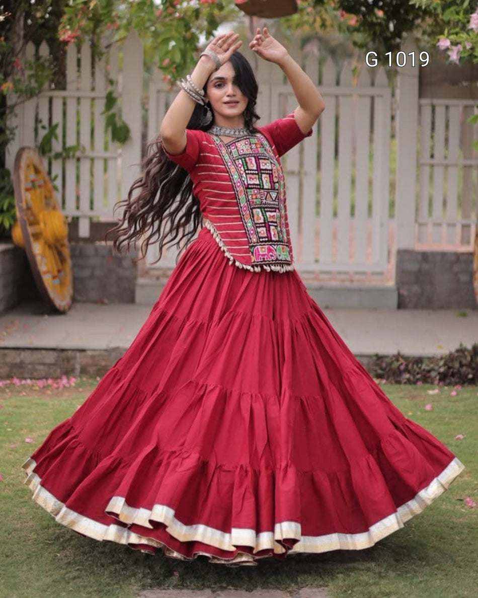 A woman wearing a traditional red Anarkali gown with a heavily embroidered neckline and a tiered flared skirt, posing outdoors in front of a white picket fence.