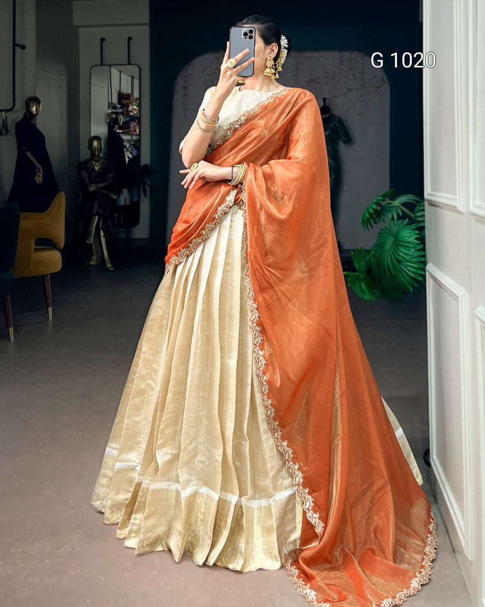 A woman takes a mirror selfie wearing a traditional Indian saree with a cream-colored flared skirt and a draped orange dupatta with gold border detailing. The background is a dark studio space with mannequins and a yellow chair.