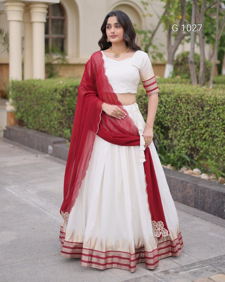 A woman modeling a traditional Indian outfit consisting of a white flared skirt (lehenga), a white sleeveless top (choli), and a deep red dupatta, standing outdoors in front of a building with columns and green hedges. The text 'G 1027' is visible in the top right corner.