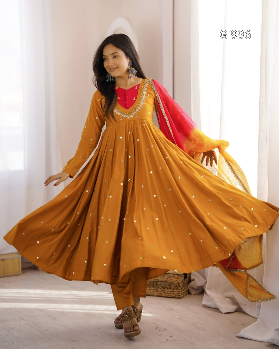 A woman modeling a traditional Indian outfit consisting of a mustard yellow Anarkali kurta with silver embroidery, matching trousers, a bright red inner neckline, and a red and yellow dupatta, captured in a brightly lit indoor setting.