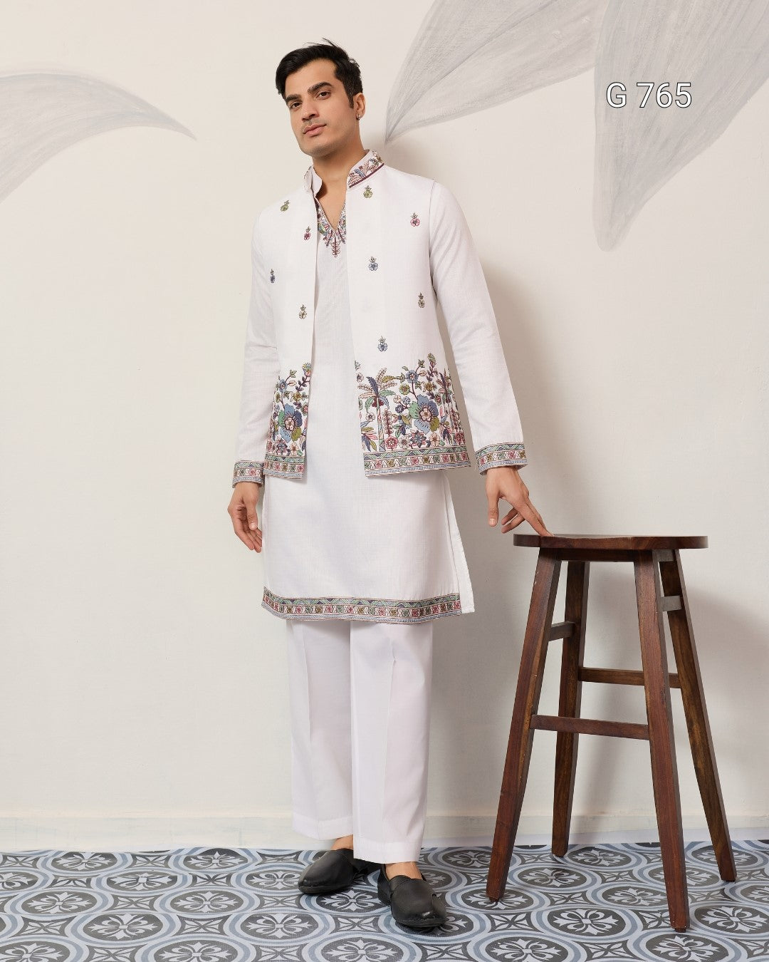 Man wearing a traditional white embroidered outfit standing next to a wooden stool against a white wall.