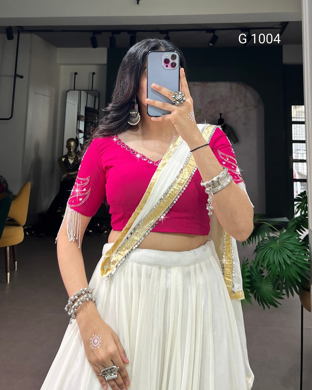 A woman takes a mirror selfie wearing a bright pink short-sleeve crop blouse with silver embellishments, paired with a white pleated skirt and a white sash with gold and silver borders. She is accessorized with silver jewelry, including a large ring, earrings, and bracelets.