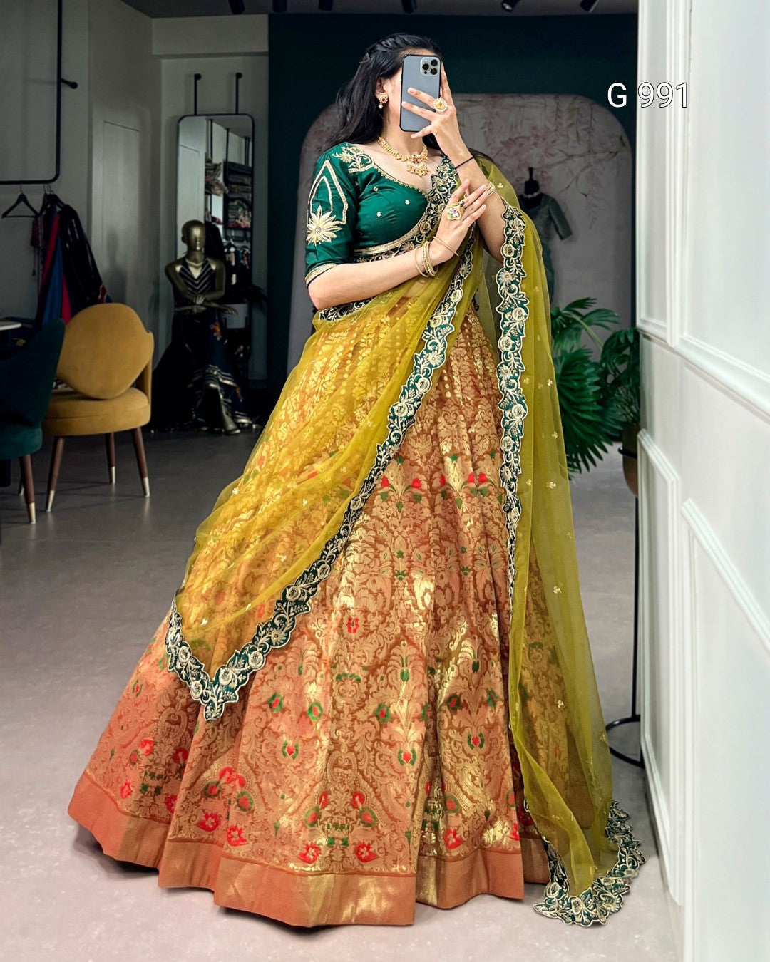 A woman wearing a traditional Indian lehenga choli dress in a mirror selfie. The outfit features a deep green embroidered blouse with gold details, a golden-yellow embroidered skirt, and a matching sheer yellow dupatta with green and gold borders.