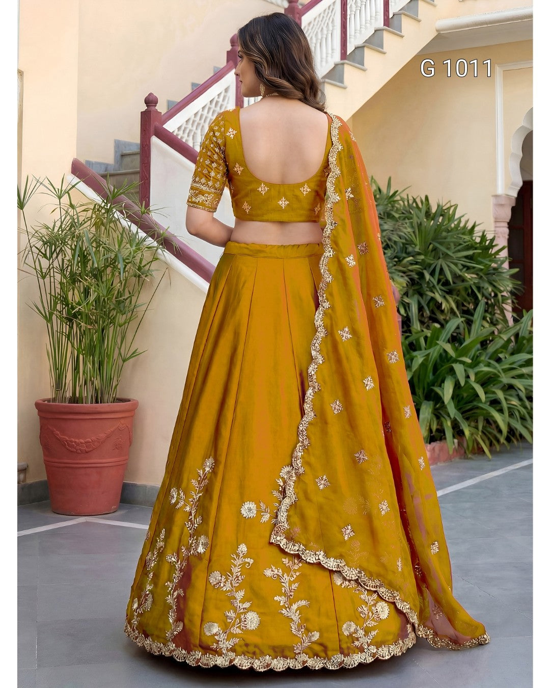 A woman wearing a mustard yellow lehenga choli with white floral embroidery and a matching dupatta, photographed from the back in an outdoor courtyard setting.