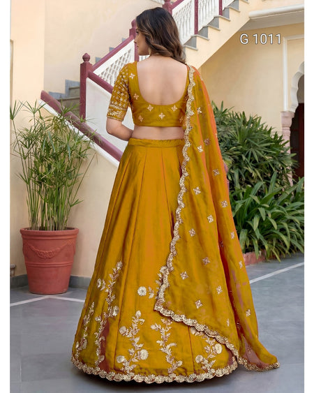 A woman wearing a mustard yellow lehenga choli with white floral embroidery and a matching dupatta, photographed from the back in an outdoor courtyard setting.