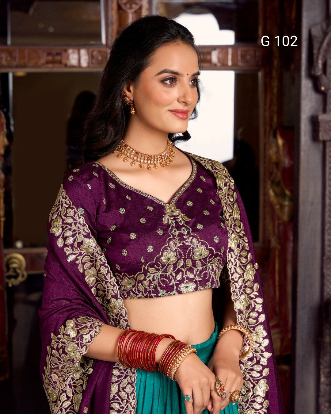 Woman wearing a traditional purple and green saree with gold embroidery, posing indoors.