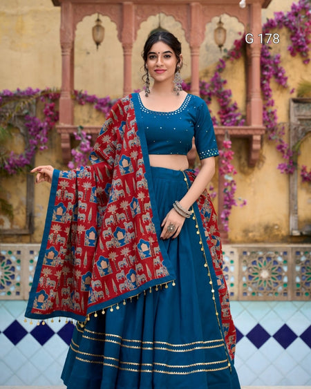 Woman in traditional blue outfit with red and gold dupatta against a decorative wall.