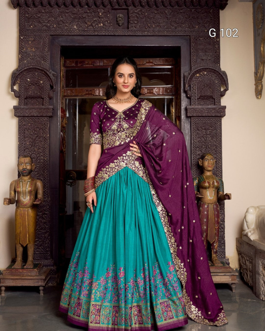 Woman wearing a purple and teal embroidered saree in an indoor setting with decorative elements.