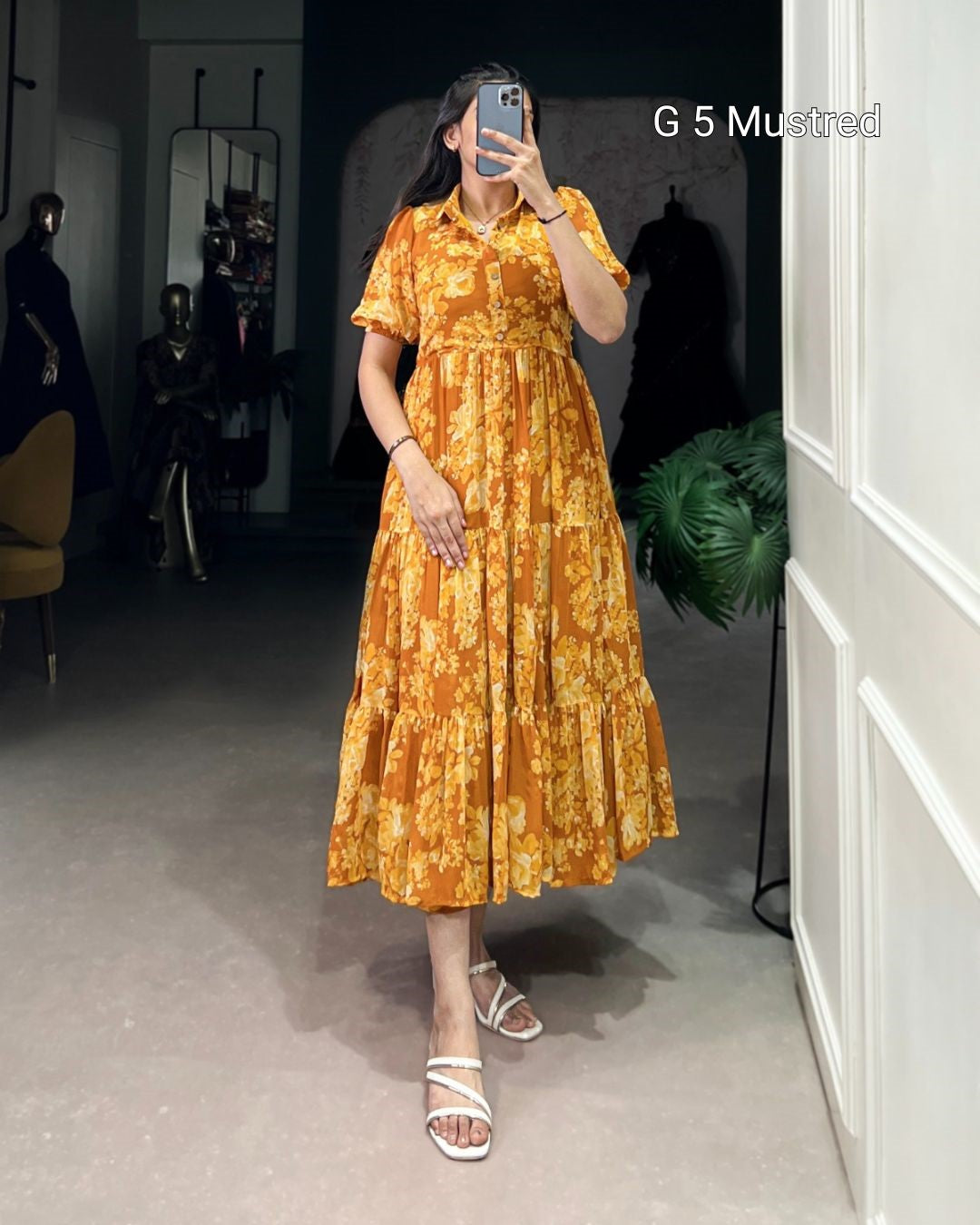 Woman taking a mirror selfie wearing an orange floral dress in a room with white walls and a plant.