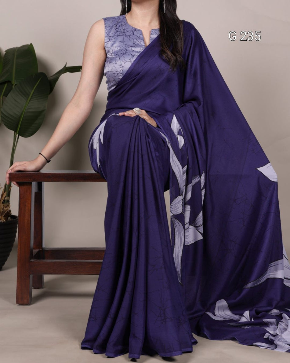 Woman wearing a purple saree with a floral pattern, sitting on a wooden stool.