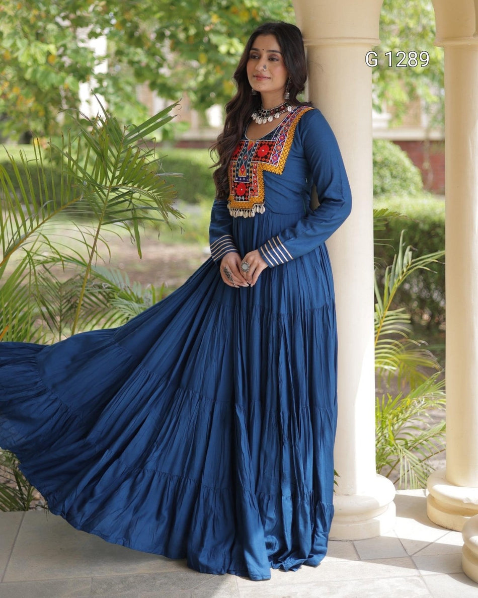 A woman wearing a long blue maxi dress with colorful embroidered details on the chest and striped cuffs, standing outdoors next to a white pillar with green plants in the background.
