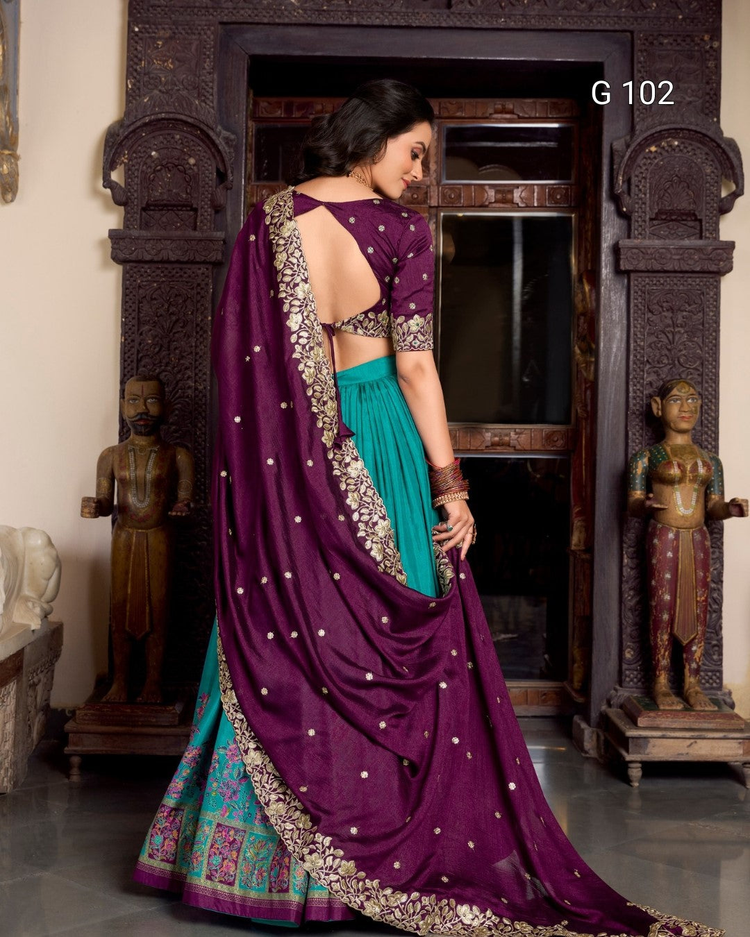 Woman in a purple and teal saree with gold embroidery standing in front of an ornate fireplace.
