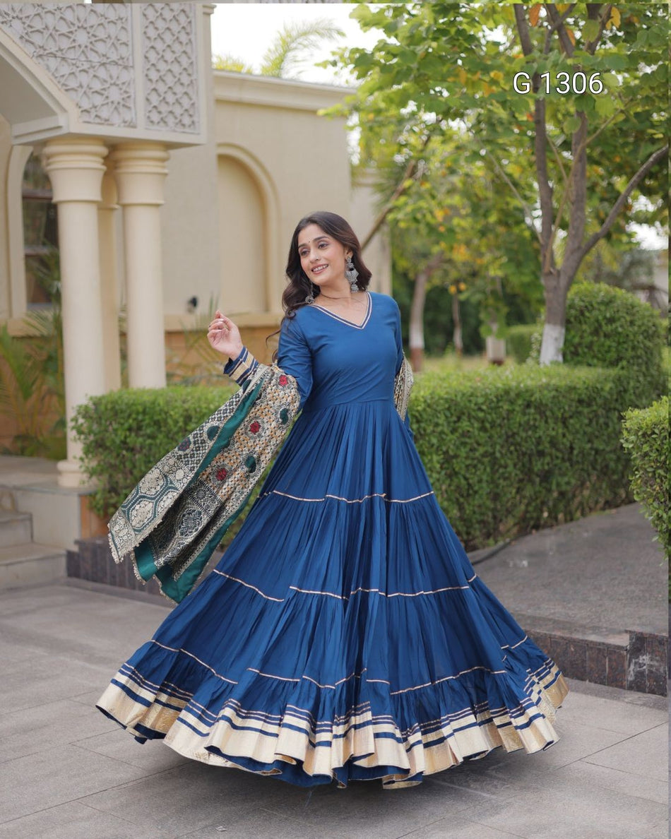 A woman outdoors wearing a blue tiered Anarkali gown with a matching dupatta featuring green, gold, and red patterns. The background shows a beige building with columns and green hedges.