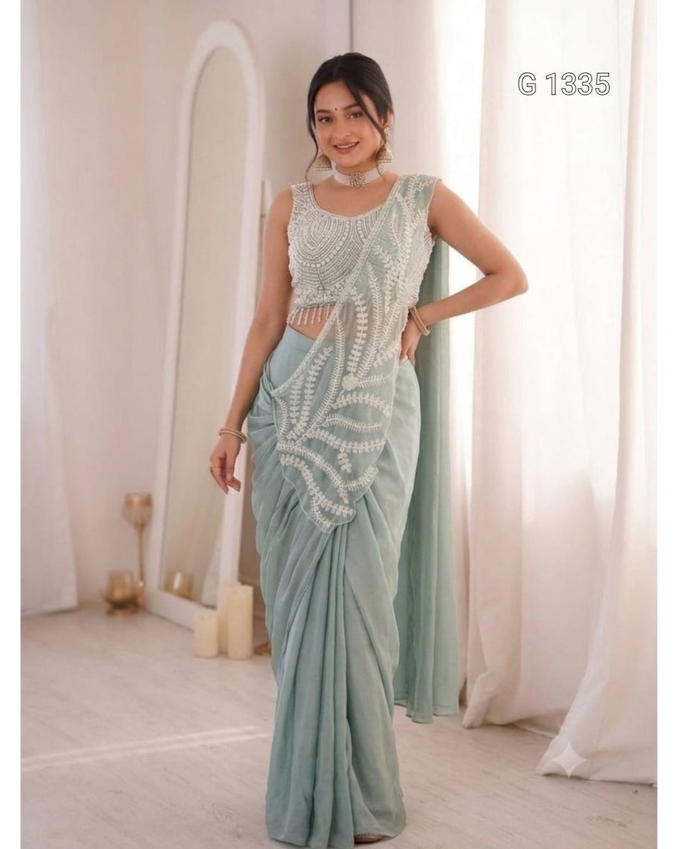 A woman wearing an elegant mint green saree with intricate white embroidery and a matching sleeveless blouse.
She is styled with traditional jewelry, standing gracefully in a softly lit, minimal indoor setting.