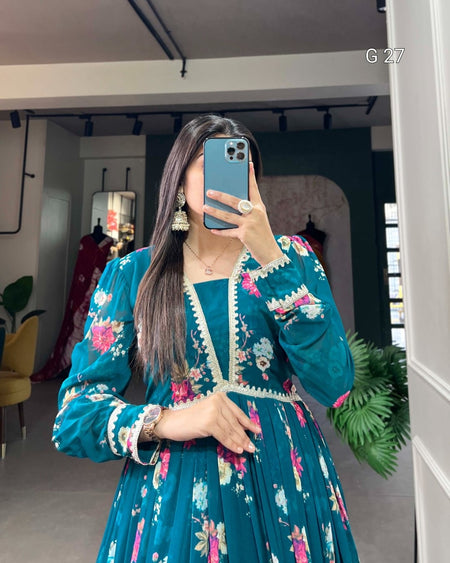Woman taking a mirror selfie wearing a teal embroidered dress in a room with plants and furniture.