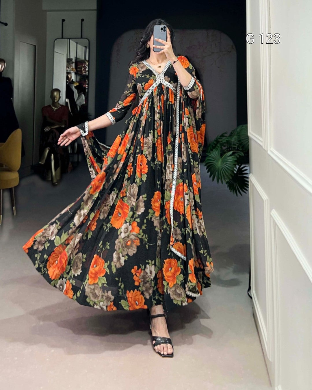 Woman in a floral dress taking a mirror selfie in a room with a white wall and plant.