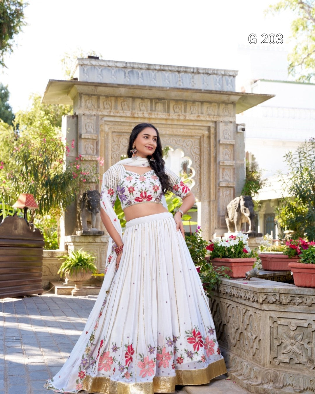 Woman in a white embroidered lehenga standing in front of a decorative stone structure with plants.