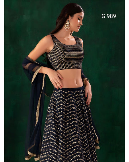 A woman modeling a black and gold embroidered traditional outfit with a sleeveless embellished crop top, patterned skirt, and draped dupatta against a dark green studio background. The text 'G 989' appears in the top right.