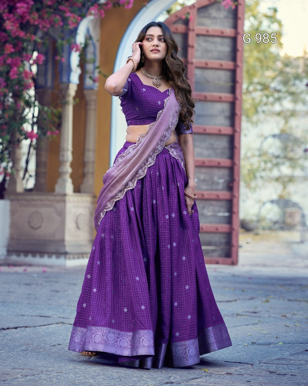 A woman modeling a purple traditional Indian outfit with a matching skirt, blouse, and dupatta in front of an outdoor architectural background with arches and wooden railings.