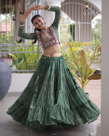 A woman modeling a dark green embroidered traditional Indian outfit with a long flared skirt and a patterned crop top, posing outdoors on a patio with plants in the background.