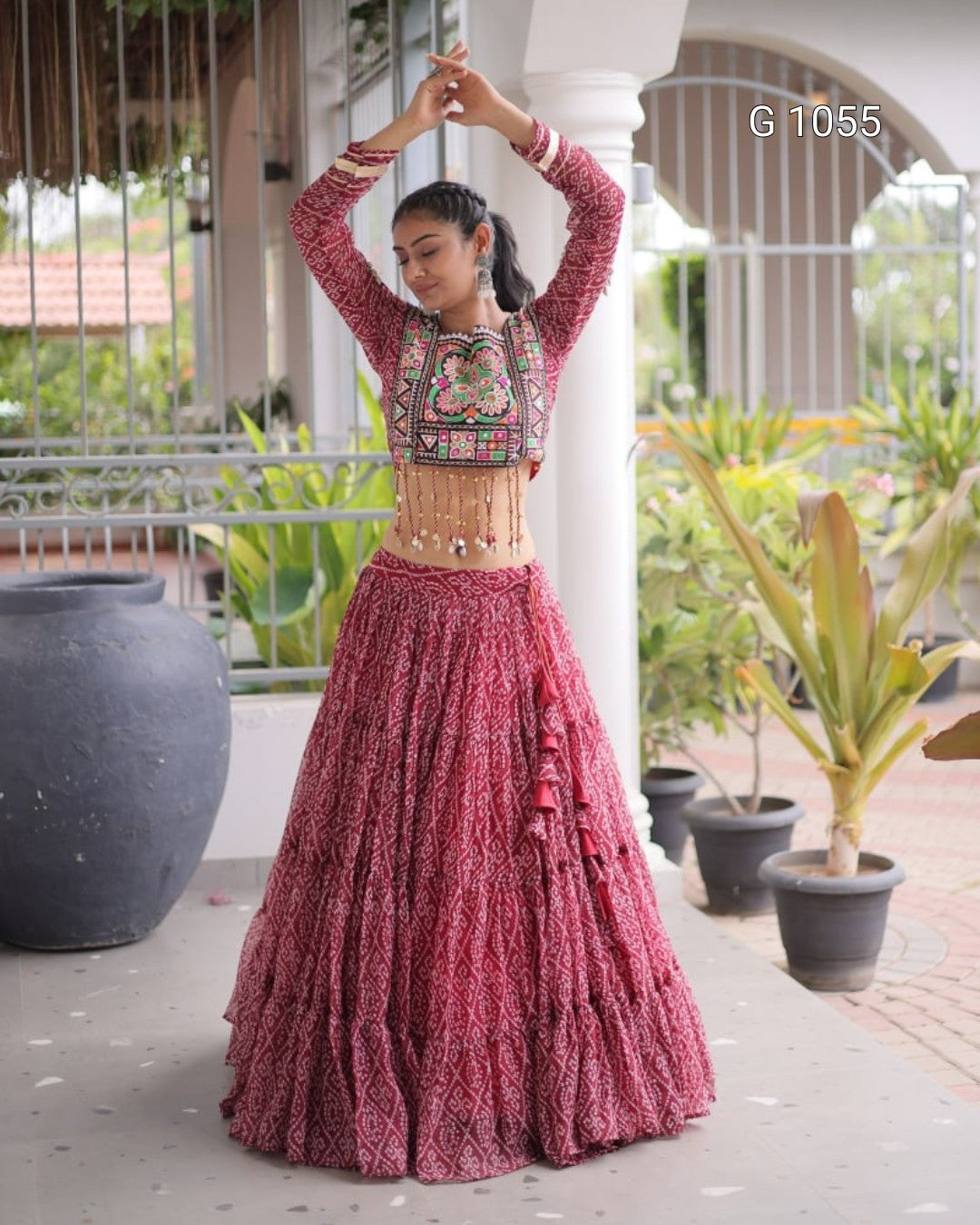 A woman modeling a traditional Indian outfit consisting of a long-sleeved embroidered crop top and a matching tiered flared skirt, in a dusty rose/magenta color, set against a residential porch background with potted plants.