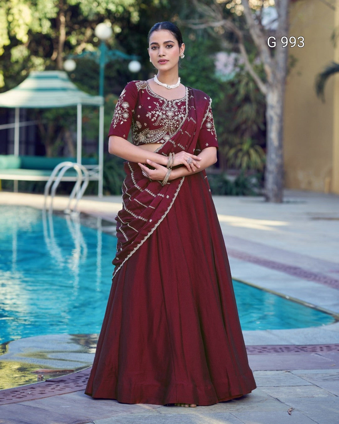 A woman stands outdoors by a swimming pool wearing a long maroon lehenga choli with gold embroidery. The image includes the text 'G 993' in the upper right corner.