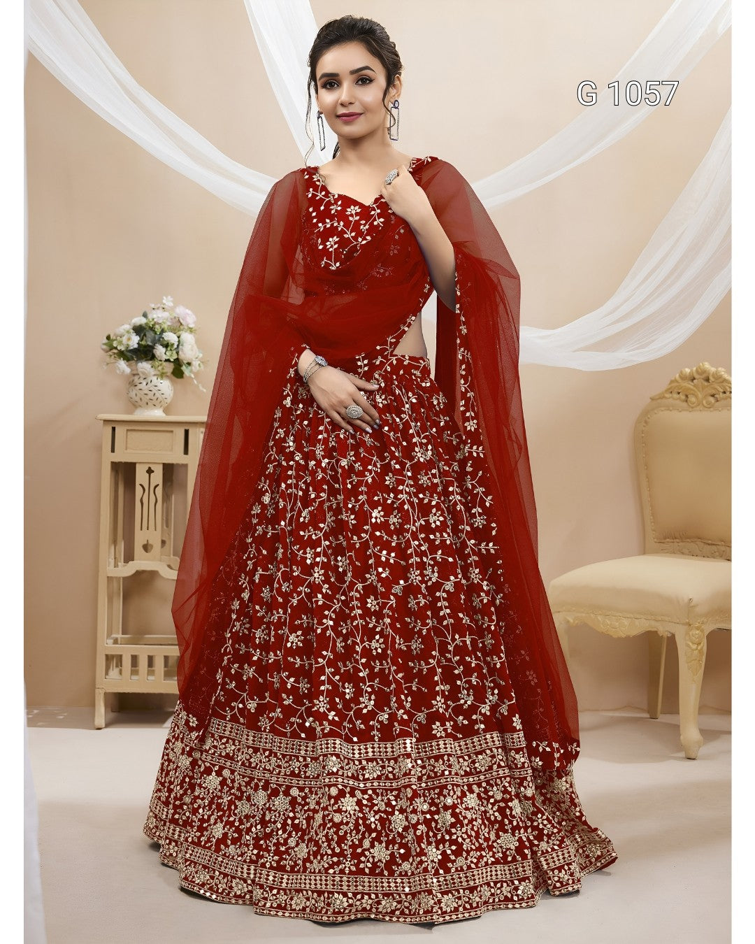 A woman models a traditional red lehenga choli set with intricate white floral embroidery and a sheer red dupatta, posed in an indoor studio setting with draped white fabric and furniture.