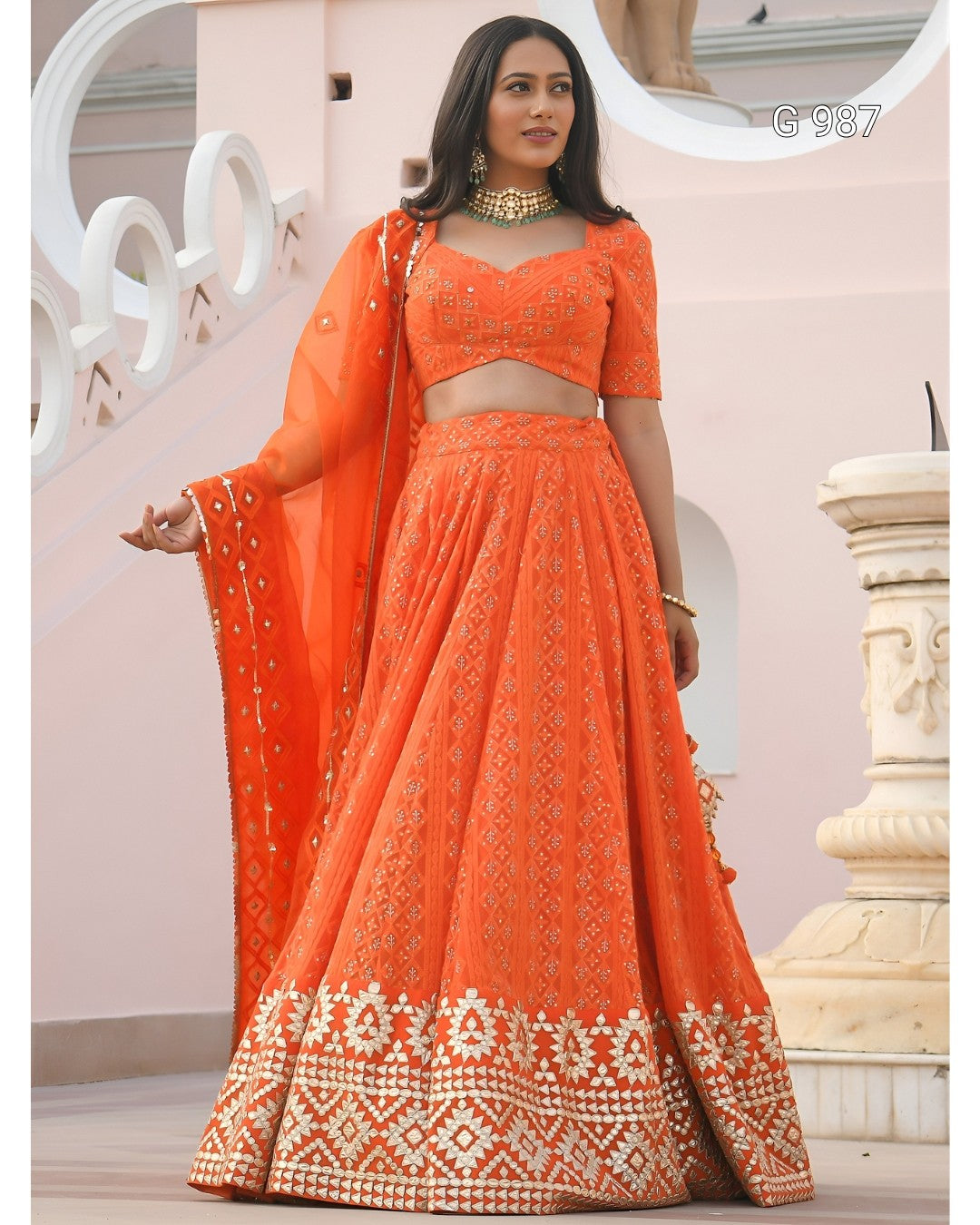 An orange embroidered Indian ethnic wear set consisting of a flared lehenga skirt, a matching crop top, and a sheer dupatta, modeled on a woman in front of a light pink architectural background.