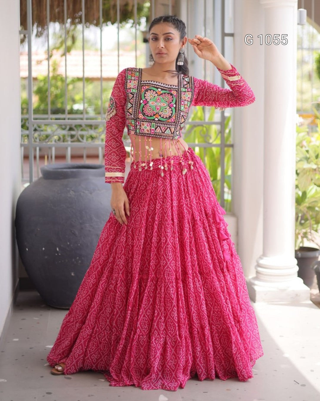 A woman models a vibrant pink embroidered traditional Indian outfit, featuring a heavily embellished floral-printed long-sleeve blouse and a matching voluminous flared skirt, set against a white-pillared outdoor corridor.