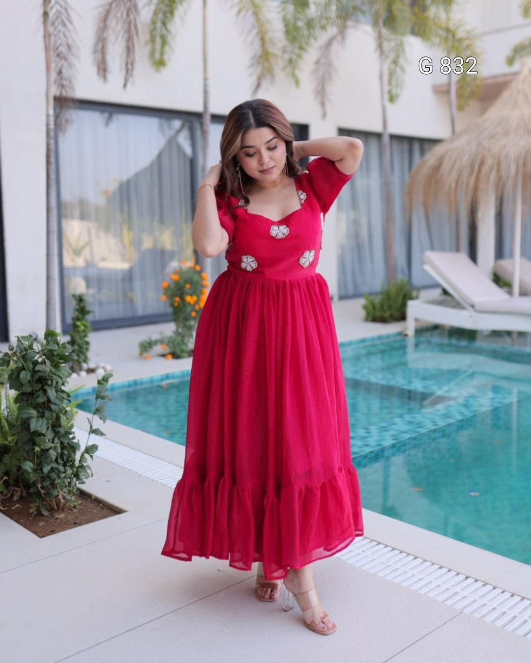 Woman in a red dress standing by a pool with plants and lounge chairs in the background.