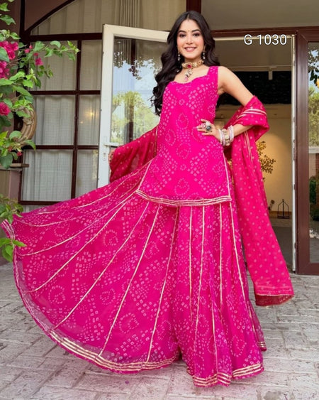 A woman modeling a bright pink Indian ethnic wear set, featuring a sleeveless kurta with bandhani tie-dye pattern, matching wide-leg flared pants, and a dupatta, posed in front of a doorway.