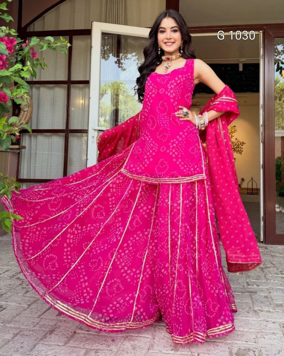 A woman modeling a bright pink Indian ethnic wear set, featuring a sleeveless kurta with bandhani tie-dye pattern, matching wide-leg flared pants, and a dupatta, posed in front of a doorway.