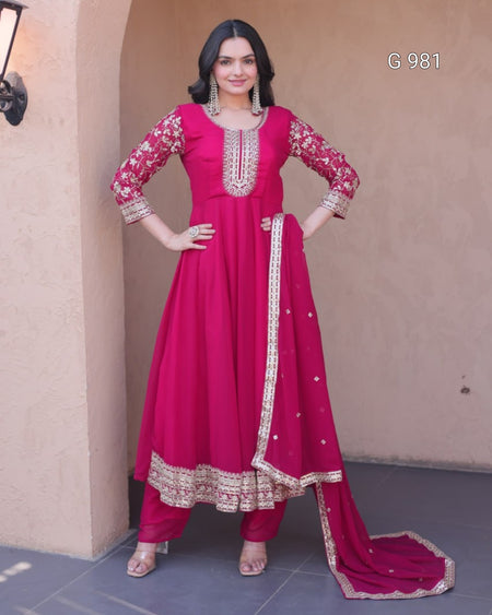 A woman modeling a bright magenta pink traditional Indian salwar kameez suit with gold embroidery, standing against a beige stucco wall. The set includes a long kurta with ornate gold embroidery on the neckline, sleeves, and hem, matching trousers, and a coordinating dupatta with a gold-trimmed border.
