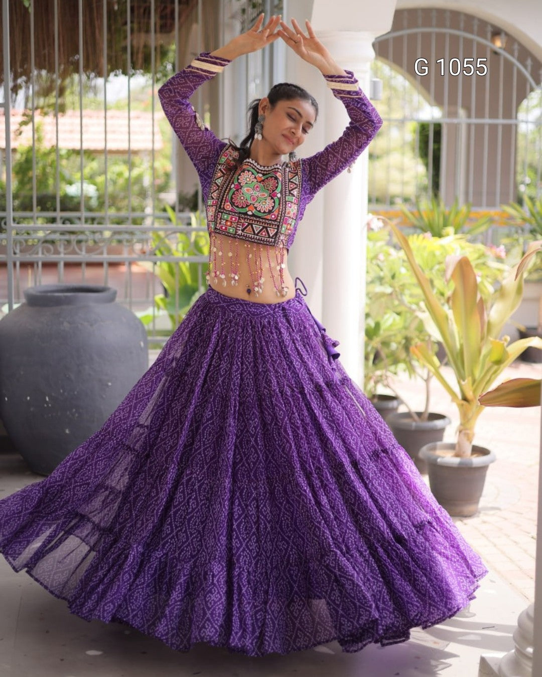 A woman modeling a purple embroidered ethnic lehenga choli set, twirling in a courtyard. The outfit features a purple lace skirt and a matching embroidered crop top with multicolor geometric patterns on the chest.