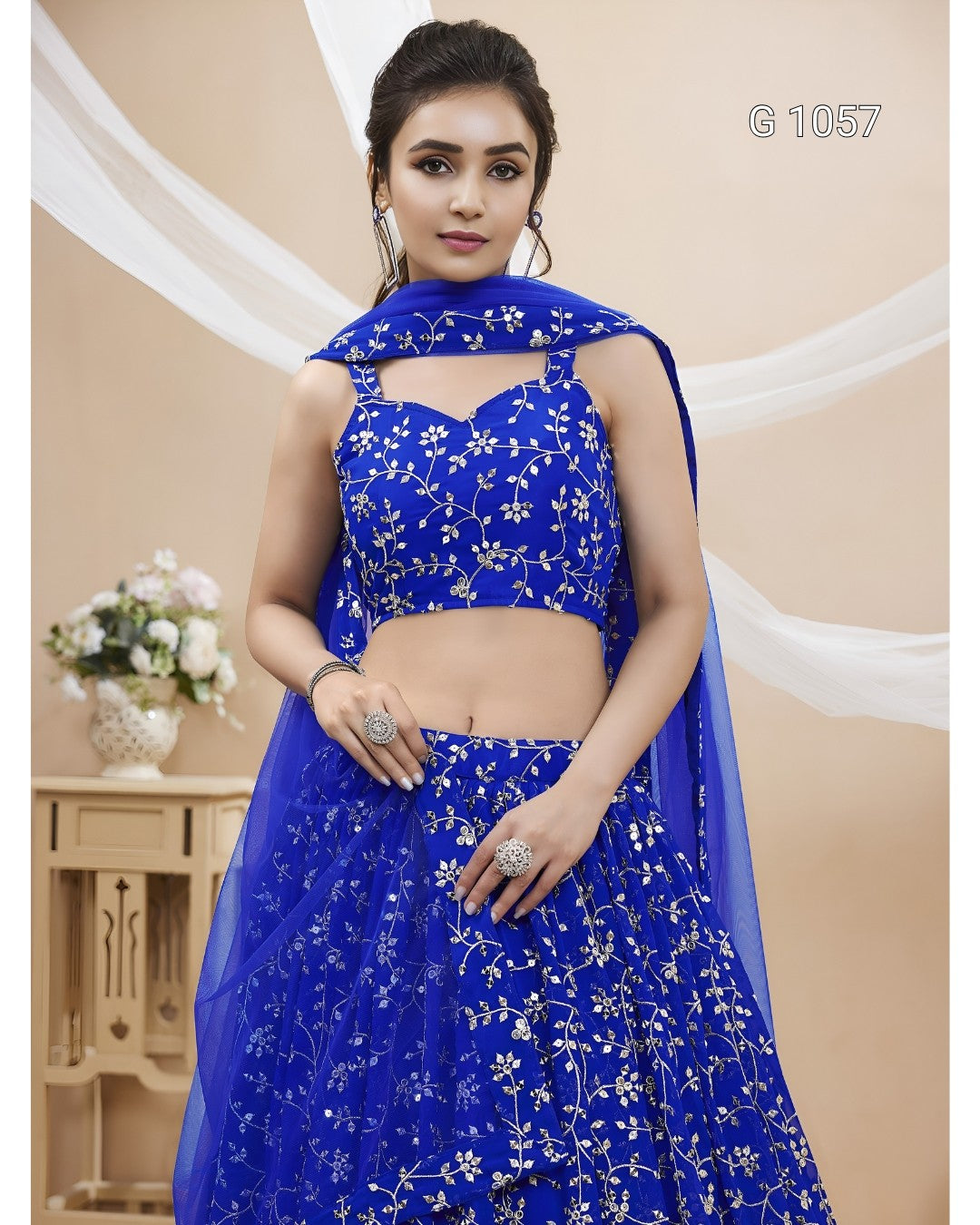 A woman modeling a royal blue lehenga choli suit with white floral embroidery, featuring a sleeveless crop top, flared skirt, and matching dupatta, set against a beige backdrop with decorative drapes.