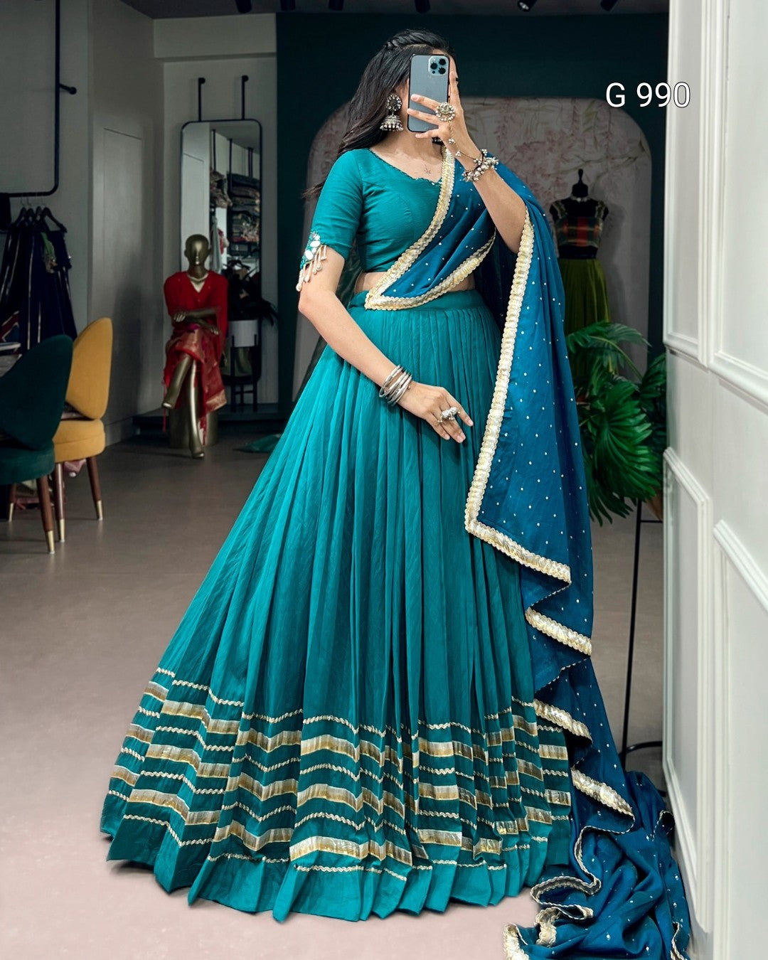 A woman takes a mirror selfie wearing a teal blue traditional Indian dress with a matching dupatta. The dress features a pleated skirt, a short sleeve with gold embroidery, and a decorative gold border at the hem.