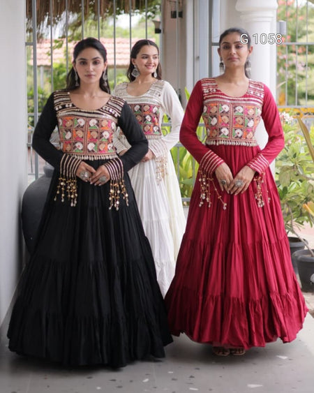 Three women modeling traditional Indian Anarkali gowns in a porch setting. The woman on the left wears a black gown with a multicolor embroidered bodice and gold beaded waist details.