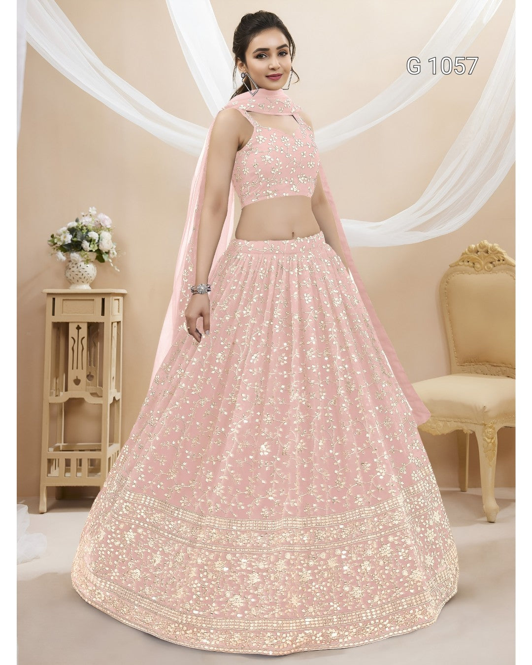 A woman models a light pink traditional Indian lehenga choli set with intricate gold embroidery, featuring a flared skirt, embroidered crop top, and a matching dupatta. The image is set in a decorated studio with beige walls, draped white fabric, and a wooden side table.
