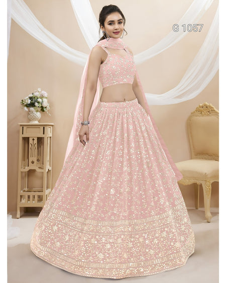 A woman models a light pink traditional Indian lehenga choli set with intricate gold embroidery, featuring a flared skirt, embroidered crop top, and a matching dupatta. The image is set in a decorated studio with beige walls, draped white fabric, and a wooden side table.