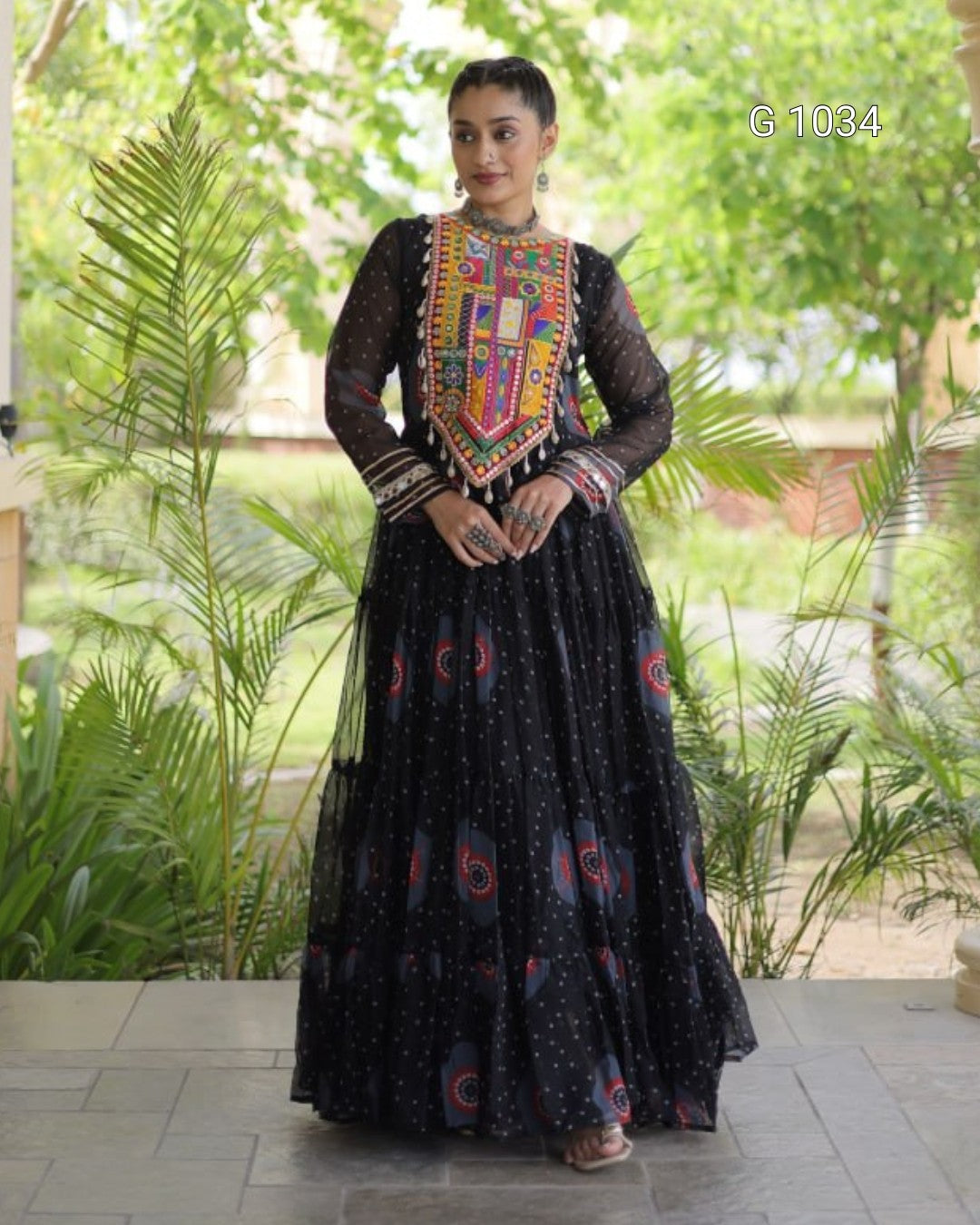 A woman wearing a black long-sleeve Anarkali gown with a heavily embroidered multicolor yoke and scattered blue and red motifs on the skirt, standing outdoors in front of green plants.