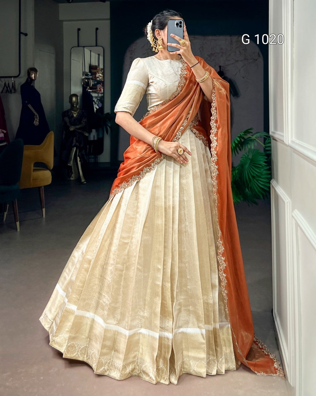 A woman takes a mirror selfie wearing a traditional Indian outfit consisting of a cream-colored flared skirt (lehenga), a cream blouse, and an orange dupatta with gold border detailing. The background is a clothing store interior.